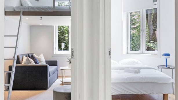 Un espacio habitable moderno y abierto con un entrepiso-dormitorio, un conjunto de sofás, una mesita y una ventana con vistas a árboles.