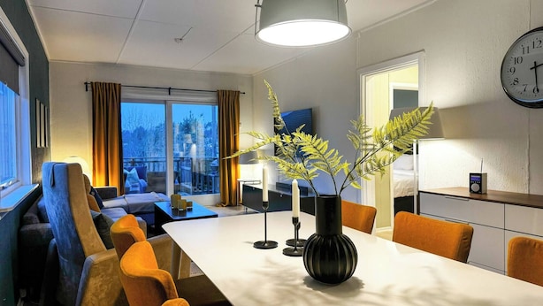 A modern hotel room with a dining area, a large window with curtains, a clock on the wall, and a plant on the table. - Cozy 2BR Apartment With Parking (Oslo)
