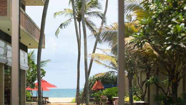 A view from a balcony overlooking a beach, with palm trees, outdoor furniture, and a red umbrella. - Ayuda Sanctuary Boutique Negombo (Negombo)