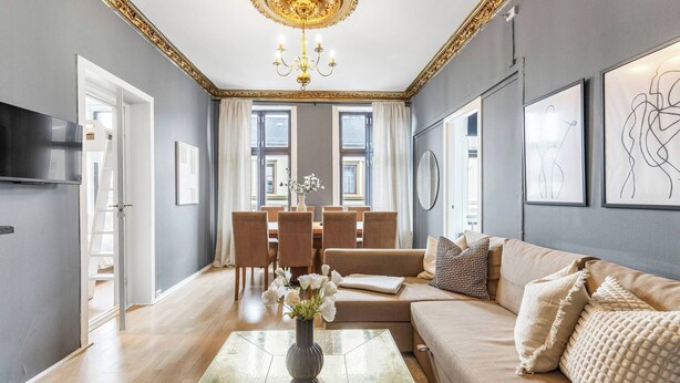 A spacious living room with a sofa, a coffee table with a vase of flowers, a dining area with chairs and a table, and a chandelier. - Big and Bright Apartment in the Center (Oslo)