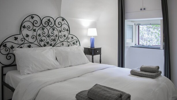 A neatly made bed with white linens and two folded towels, a bedside lamp, and a window with curtains. - Camula Inn (Catania)