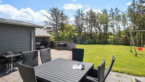 En udendørs terrasse med et bord, stole, grill og gyngestativ.