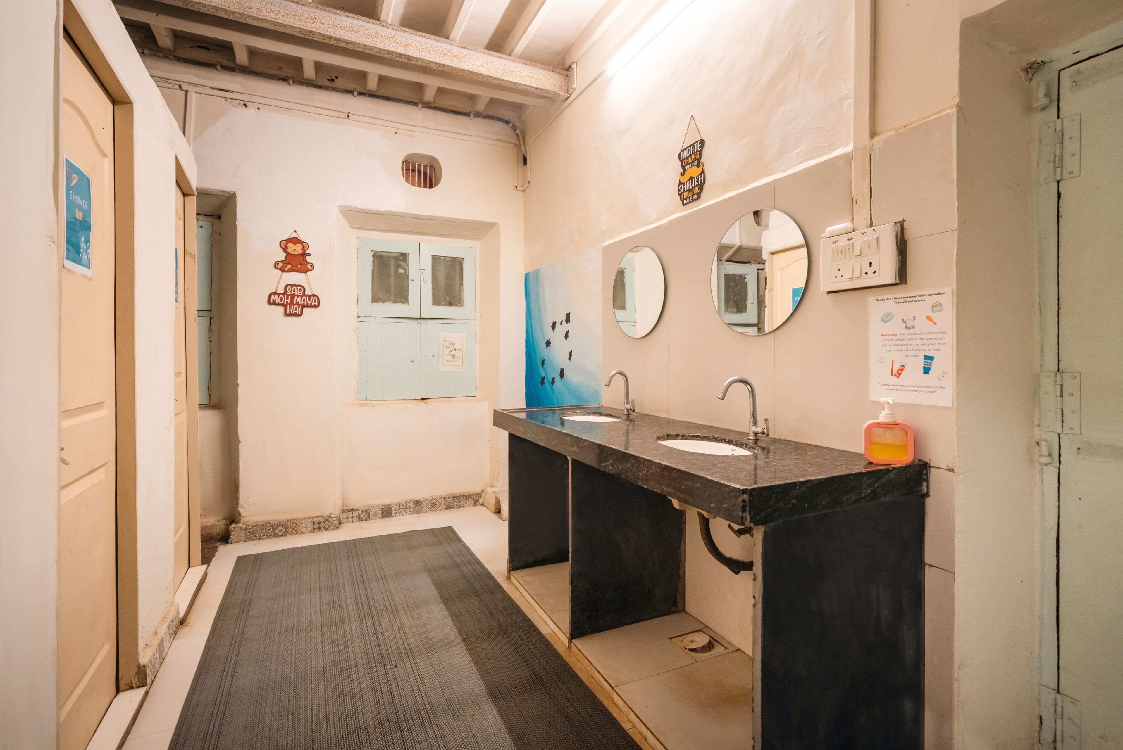 A bathroom with two sinks, a mirror, a hand sanitizer dispenser, and a door with a light switch.