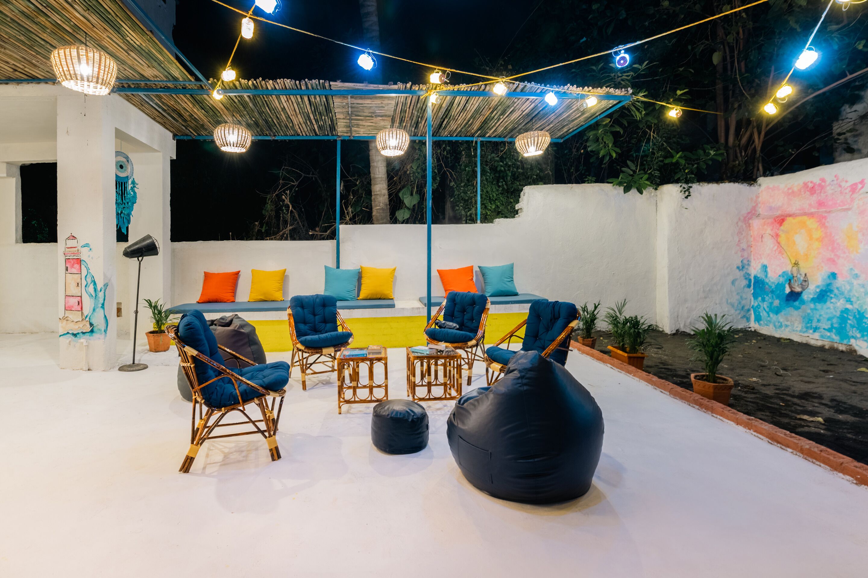 Un espace extérieur aménagé avec des chaises en osier et un pouf noir, situé sous une terrasse couverte et illuminée de guirlandes lumineuses.