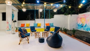 Un espace extérieur aménagé avec des chaises en osier et un pouf noir, situé sous une terrasse couverte et illuminée de guirlandes lumineuses.