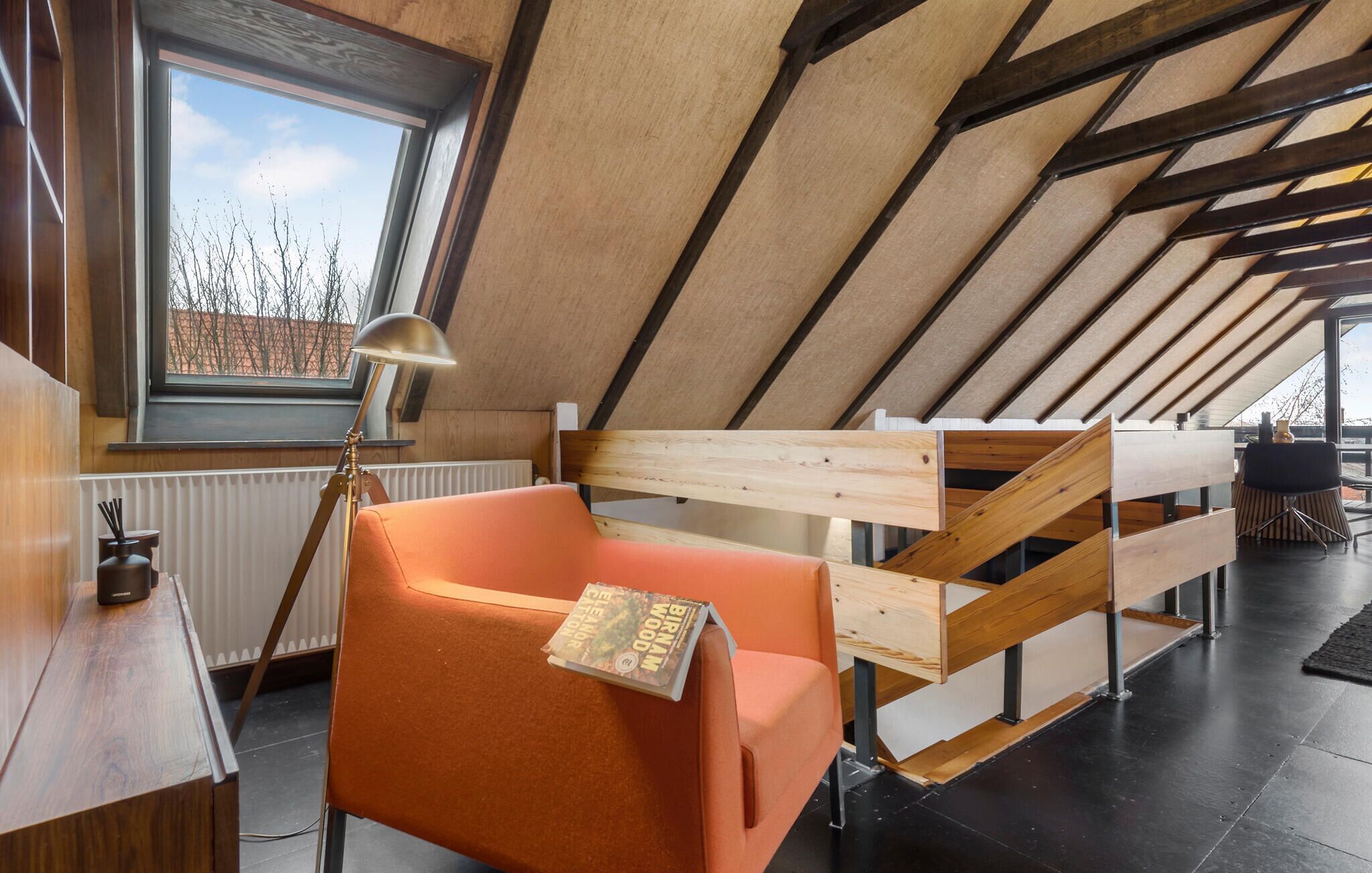 A modern loft bedroom with a sloped ceiling, a wooden headboard, a plush orange chair, a floor lamp, and a view of the outside through a large window.
