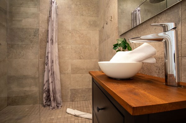 A bathroom with a marble shower wall, a wooden vanity with a white vessel sink, and a potted plant.