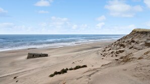 A beach with a sandy shore, a small structure, and a grassy cliff. - Lovely home in Hvide Sande with sauna (Hvide Sande)