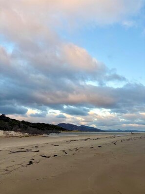 A beach with footprints leading to the water, a clear sky, and distant mountains. - Birdsong (Coles Bay)