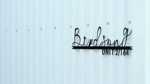 Une plaque métallique portant l’inscription « Birdsong » et « UNIT 2/164 », fixée à un mur en métal ondulé.