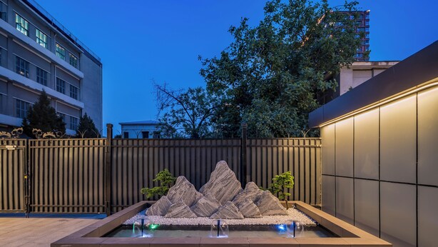 Una moderna fuente exterior con formaciones rocosas y un estanque reflectante, rodeada por una valla de madera y árboles.
