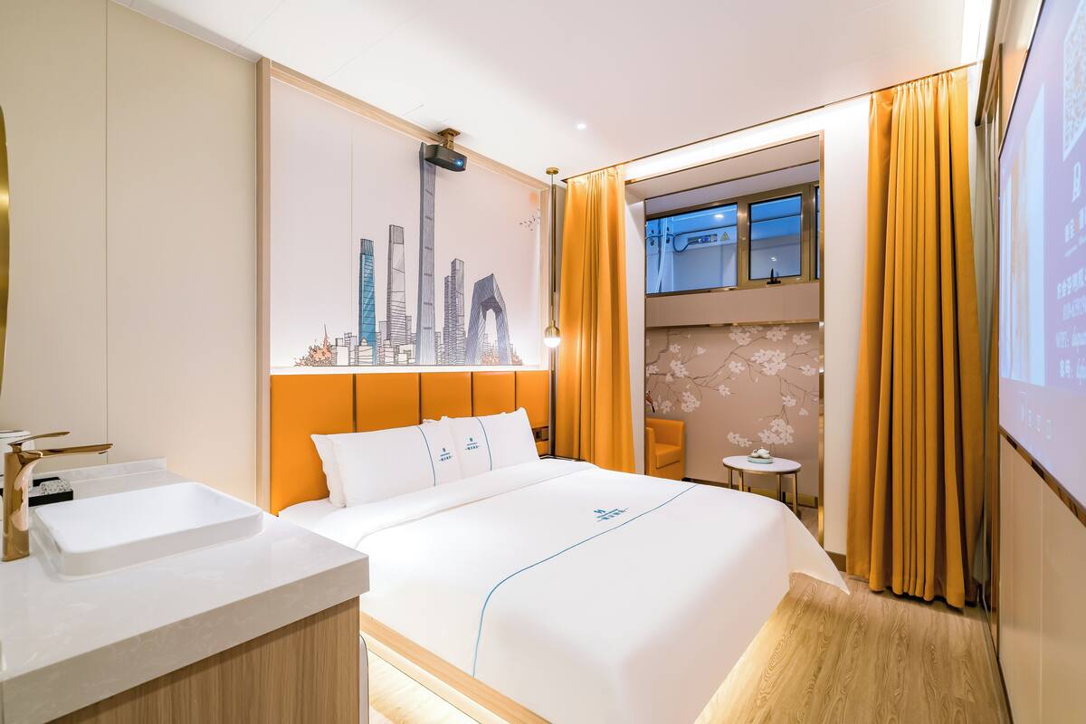 A hotel room with a large bed, a bathroom with a sink, a window with curtains, and a cityscape mural on the wall.