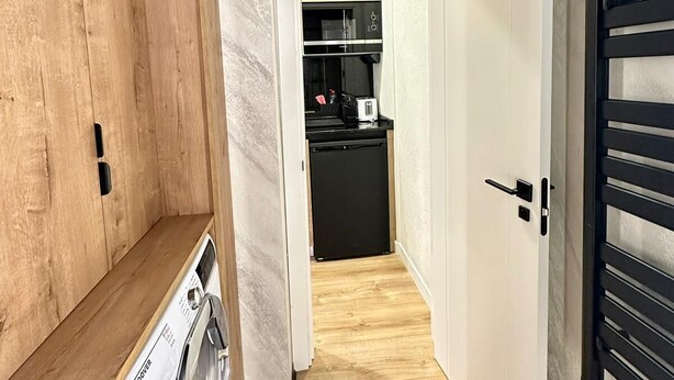 Un couloir étroit, avec une machine à laver sur la gauche, un coin cuisine avec un réfrigérateur noir et un four à micro-ondes au centre, et une porte avec une poignée noire sur la droite.