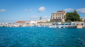 A marina with boats docked, a large building with arched windows, and a clear blue sky. - LEVANZO Standard room with small private patio (Favignana)