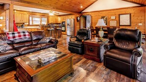 A spacious living room with a leather sofa, a recliner, and a coffee table. A kitchen with wooden cabinets and a refrigerator is visible in the background. - Escape to our cozy lakeview cabin with modern amenities, spacious decks and serene surroundinngs. (Radford)