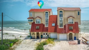 Une maison à deux étages avec un toit rouge, située sur une plage de sable, avec l’océan en toile de fond.