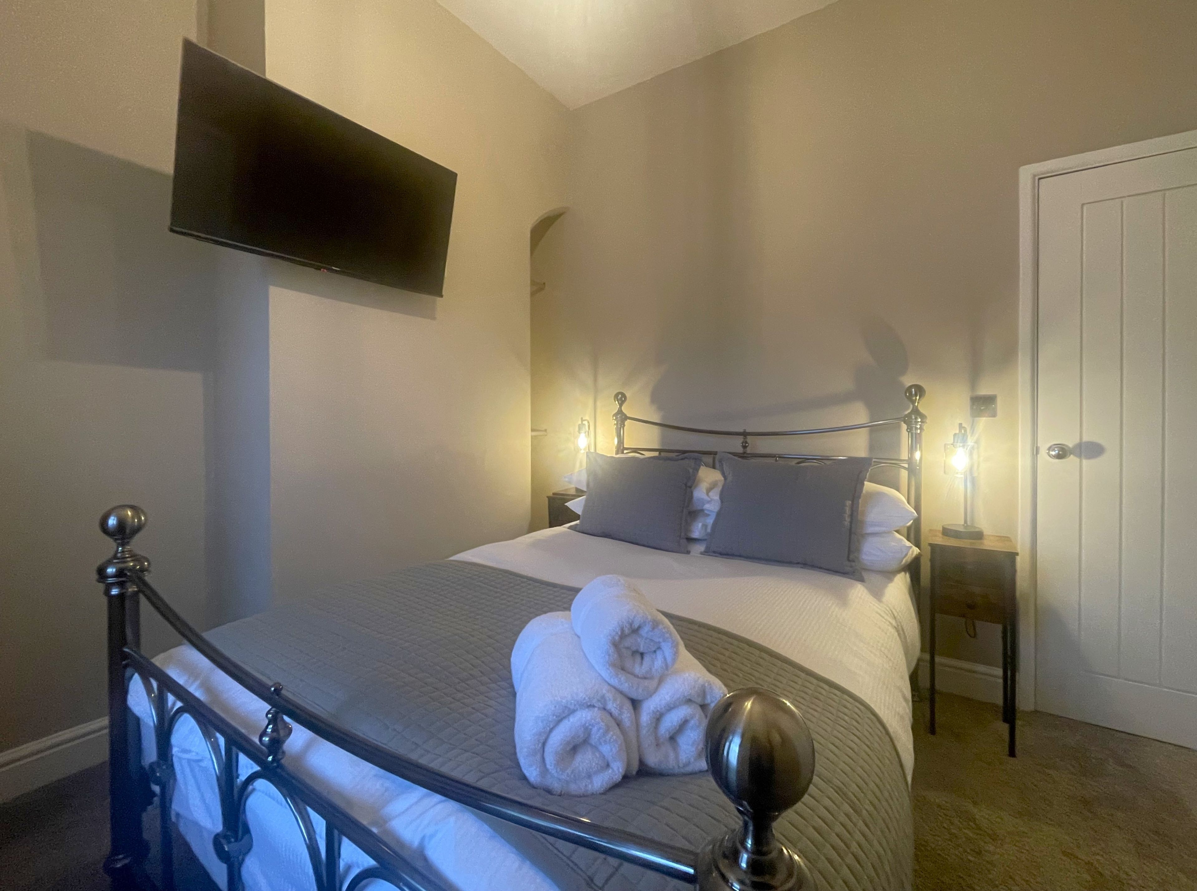 A neatly made bed with pillows and rolled towels, a bedside table with a lamp, and a flat-screen TV mounted on the wall.
