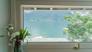A window with a view of a calm lake, a mountain in the background, and a vase with flowers on the windowsill. - Sala Comacina Villas by Rent all Como (Sala Comacina)