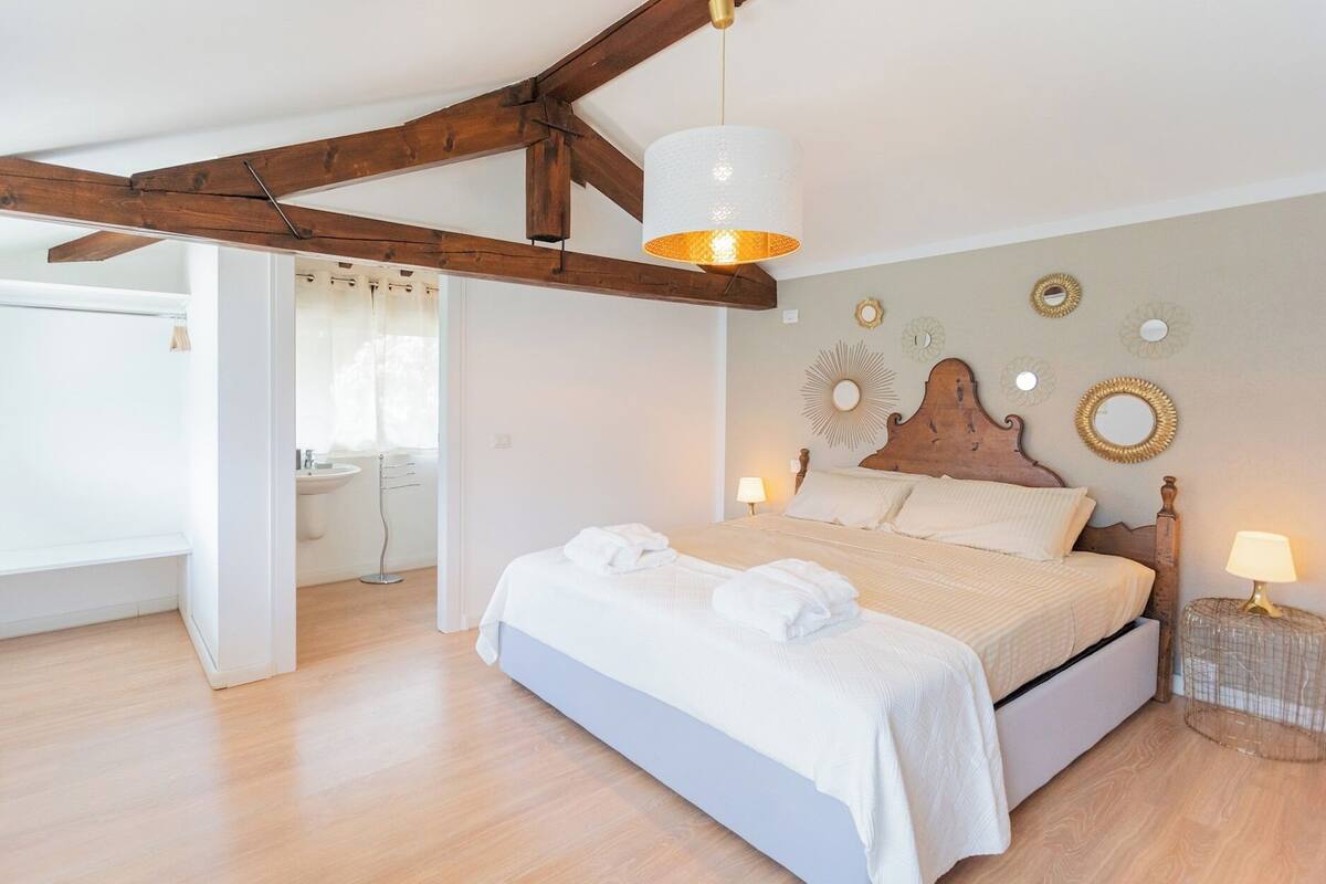A bedroom with a wooden ceiling, a bed with white linens, a bedside lamp, and wall decor.