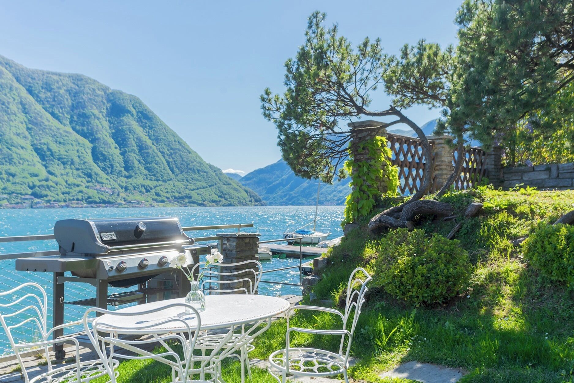 An outdoor dining area with a grill, chairs, and a view of a lake and mountains.