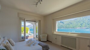A bedroom with a bed, a chair, a window with a view of a lake and mountains, and a door leading to an outdoor area. - Sala Comacina Villas by Rent all Como (Sala Comacina)