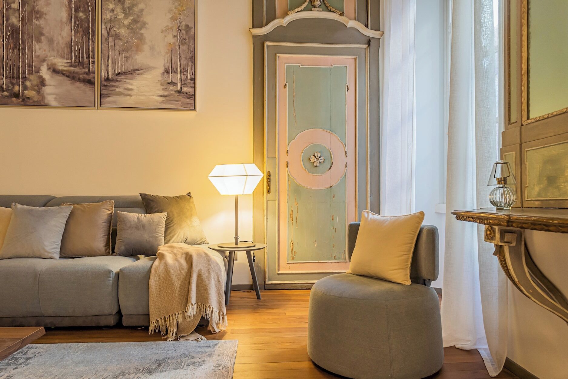 A cozy living room with a sofa, armchair, and a lamp. There's a door with a clock and a painting on the wall.