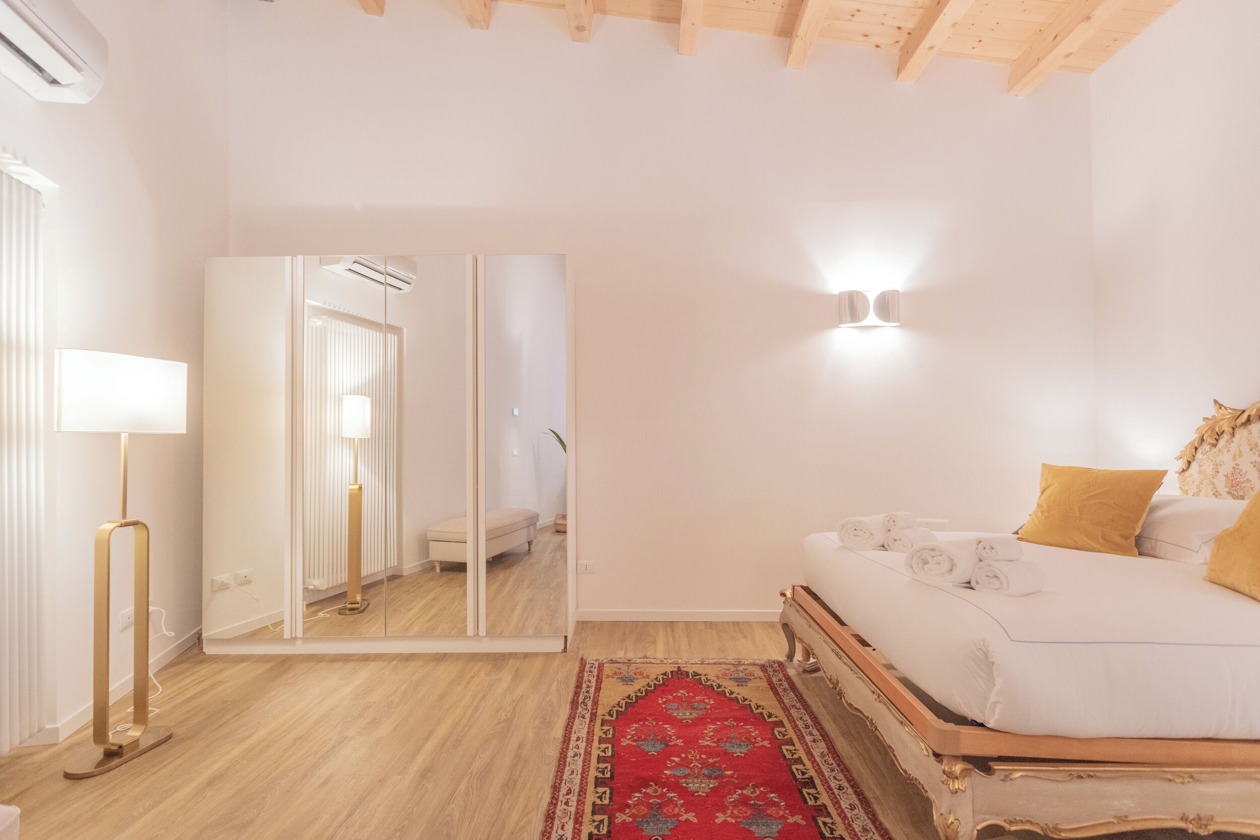 Ein Schlafzimmer mit einem Bett, einem Spiegel, einer Nachttischlampe, einem roten Teppich und einer Wandleuchte.