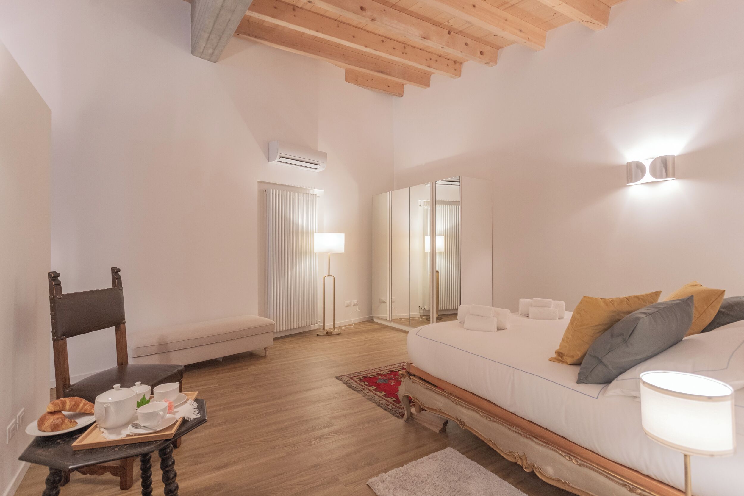 A hotel room with a bed, a chair, a small table with breakfast items, a lamp, and a mirror.