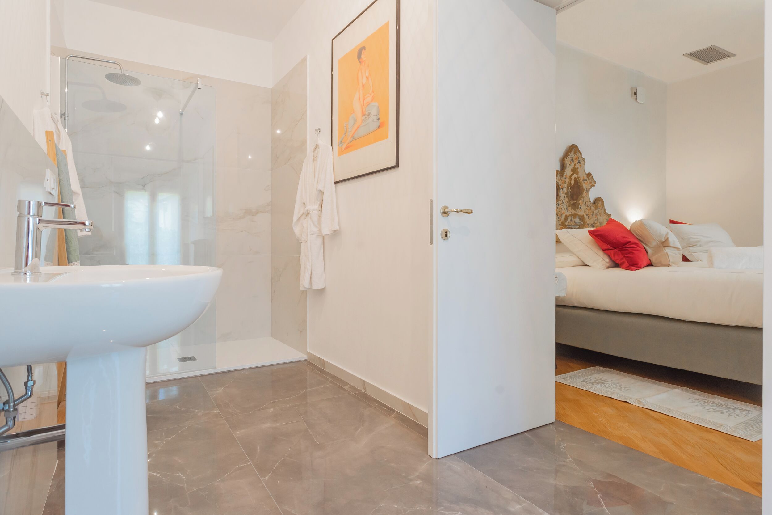 A modern bathroom with a glass shower, a white sink, and a bed with red pillows in the adjacent bedroom.