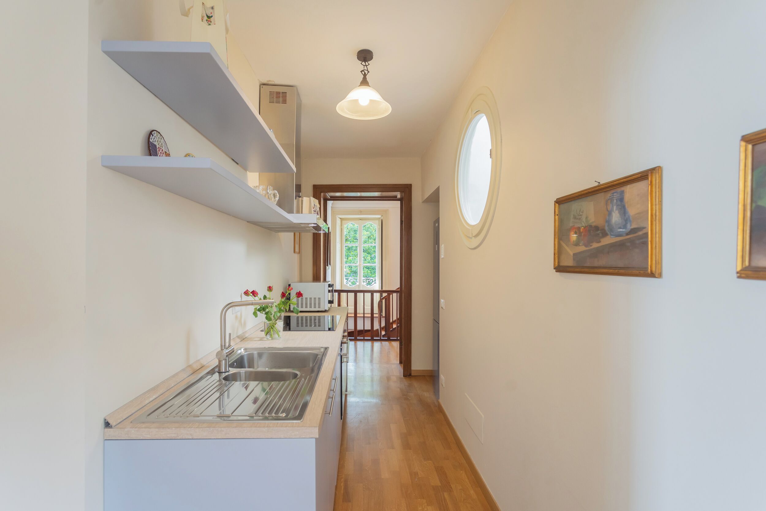 Una cocina estrecha con un lavamanos empotrado, estanterías blancas y una escalera de madera que sube a otra habitación.