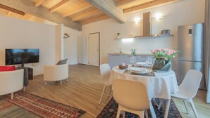 Un salon moderne avec un téléviseur, des canapés blancs, une table à manger agrémentée d’un vase de fleurs et un coin cuisine avec un réfrigérateur et des placards.
