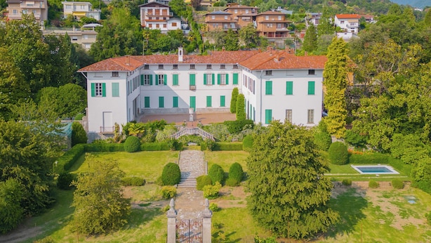 Un grand bâtiment blanc aux volets verts, entouré d’une végétation luxuriante et d’un jardin bien entretenu.