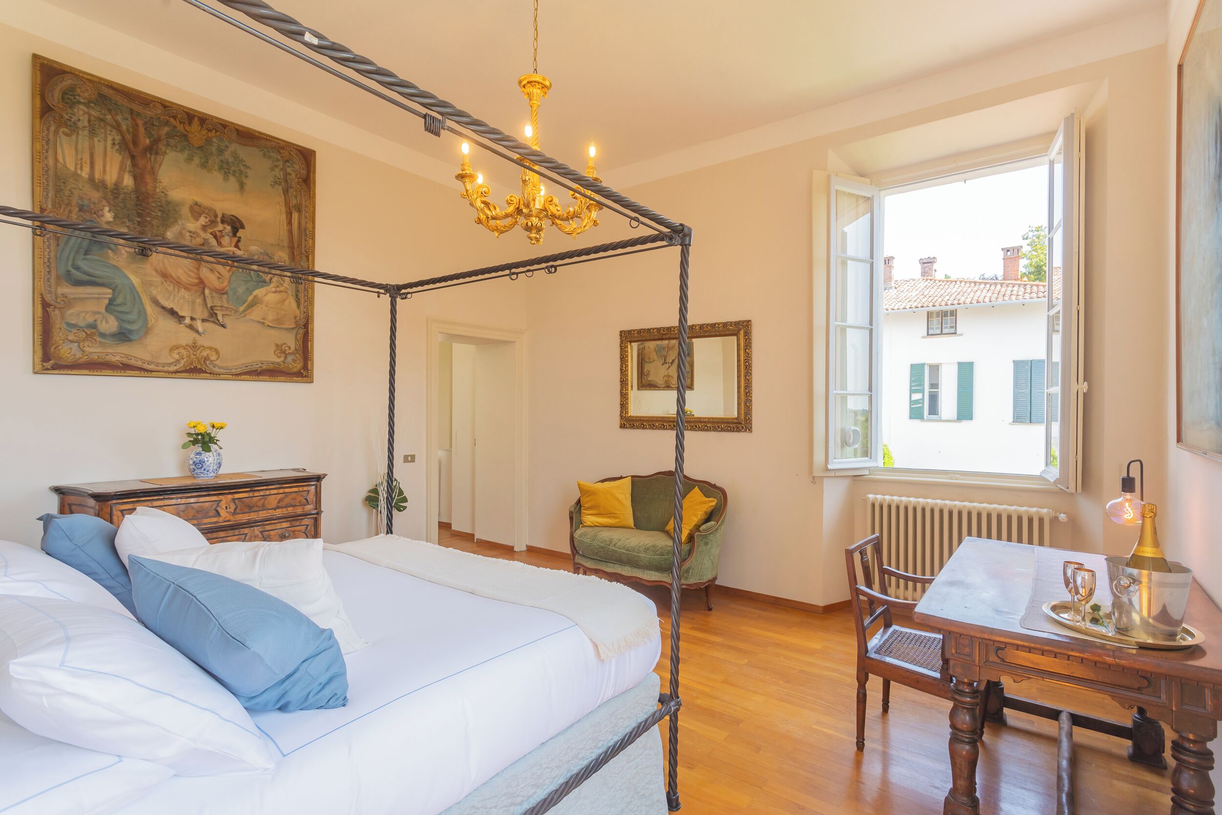 A four-poster bed with a chandelier, a dining table with chairs, a green armchair, and a painting on the wall.
