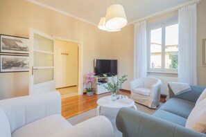 Een woonkamer met een flatscreen televisie, twee witte fauteuils, een grijze bank, een klein witte salontafel en een potplant met roze bloemen.