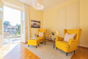 Een lichte kamer met gele stoelen, een glazen tafel en uitzicht op een huis door een geopende deur.