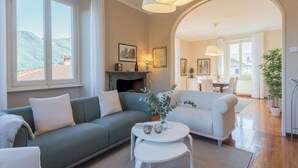 A living room with a sofa, a coffee table, and a fireplace. There are windows with a view of the mountains. - Villa Ida by Rent all Como (Tremezzina)