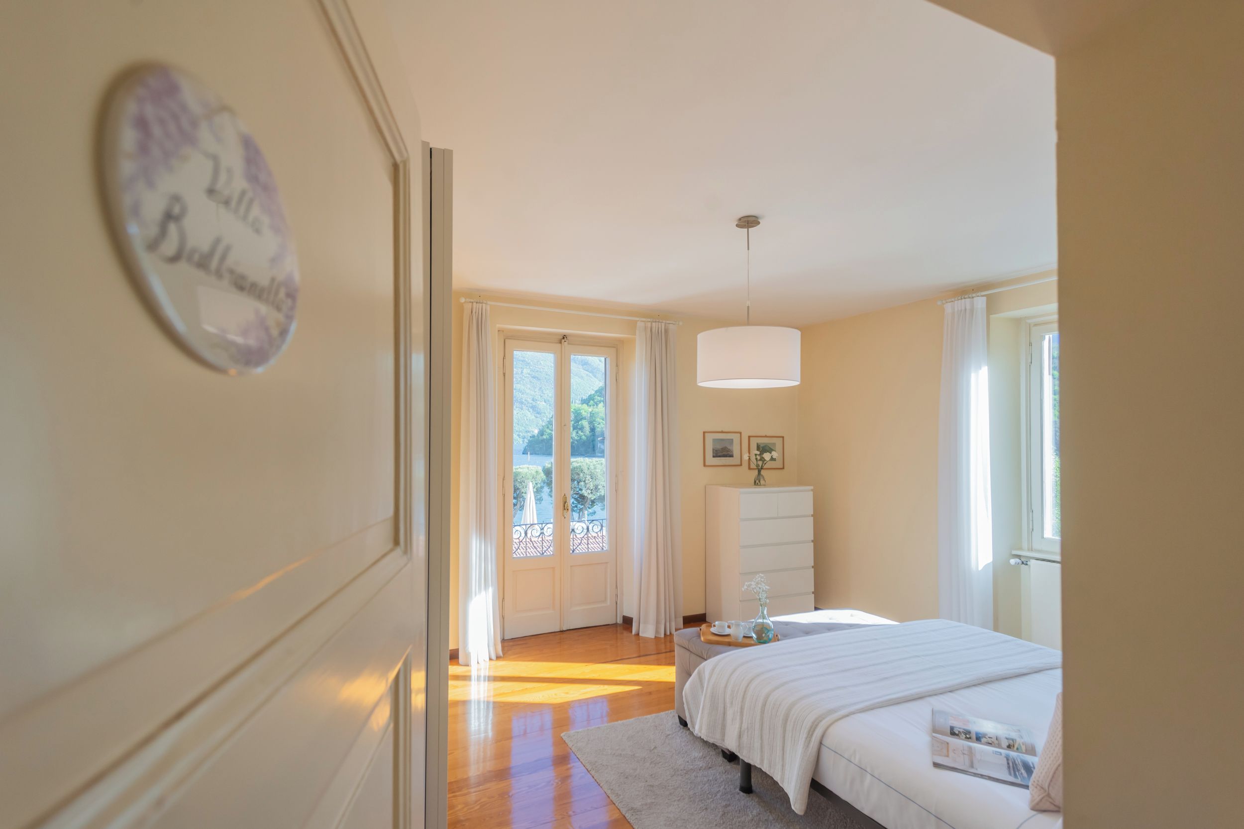 A bedroom with a bed, a bedside table, a dresser, a hanging light, and a door leading to a balcony.