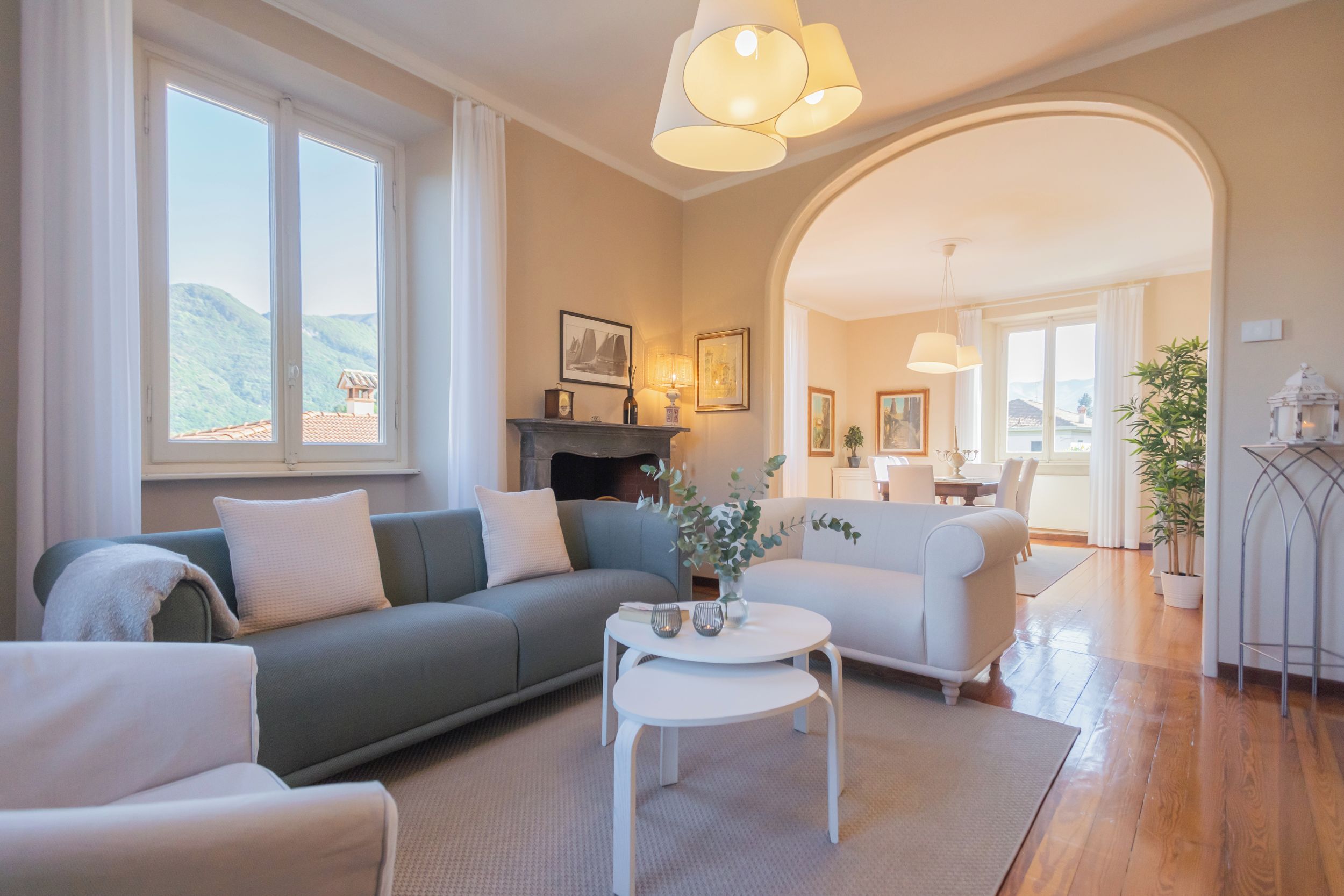 A spacious living room with a grey sofa, a white armchair, and a small white table. There is a fireplace with a mantel, framed pictures, and a large window with a view of hills.