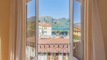 Un balcon doté d’une balustrade en fer forgé, donnant sur une ville côtière avec des bâtiments aux toits de tuiles et un paysage montagneux en arrière-plan.