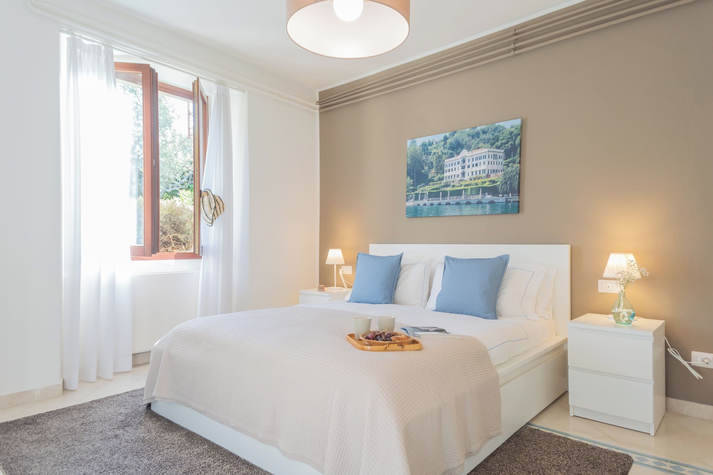 A bedroom with a large bed, a nightstand with a lamp, a window with curtains, and a painting on the wall.