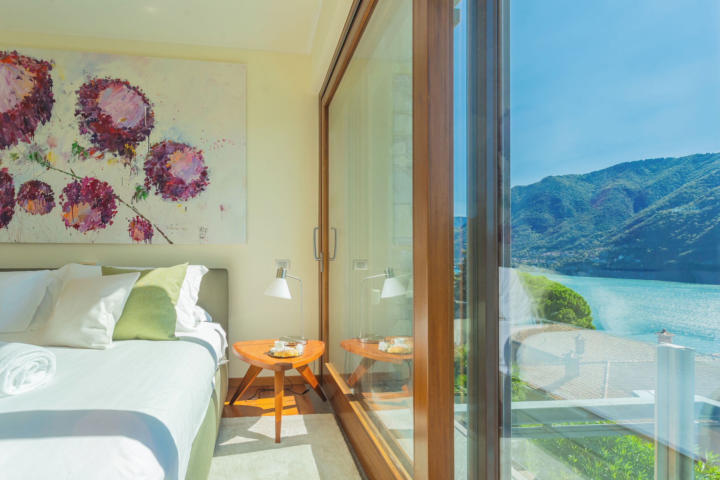 A bedroom with a bed, a painting, and a balcony with a view of a lake and mountains.