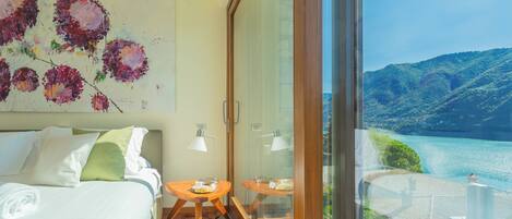 A bedroom with a bed, a painting, and a balcony with a view of a lake and mountains.