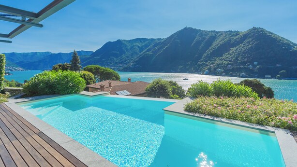 Ein Schwimmbecken mit Holzterrasse, umgeben von üppiger Vegetation und Blick auf Berge und einen See.