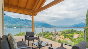 A balcony with a wooden ceiling, furnished with a sofa, chairs, and a table, overlooking a lake and mountains. - Villa Cosima in Bellagio With Private Pool by Rent all Como (Bellagio)