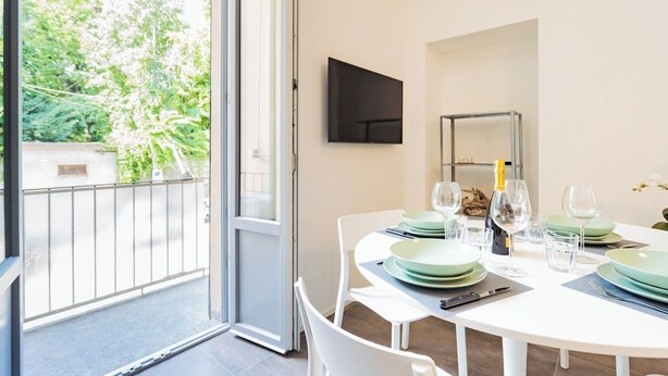 Un comedor con una mesa preparada para cuatro personas, un televisor montado en la pared y un balcón con vistas a zonas verdes.