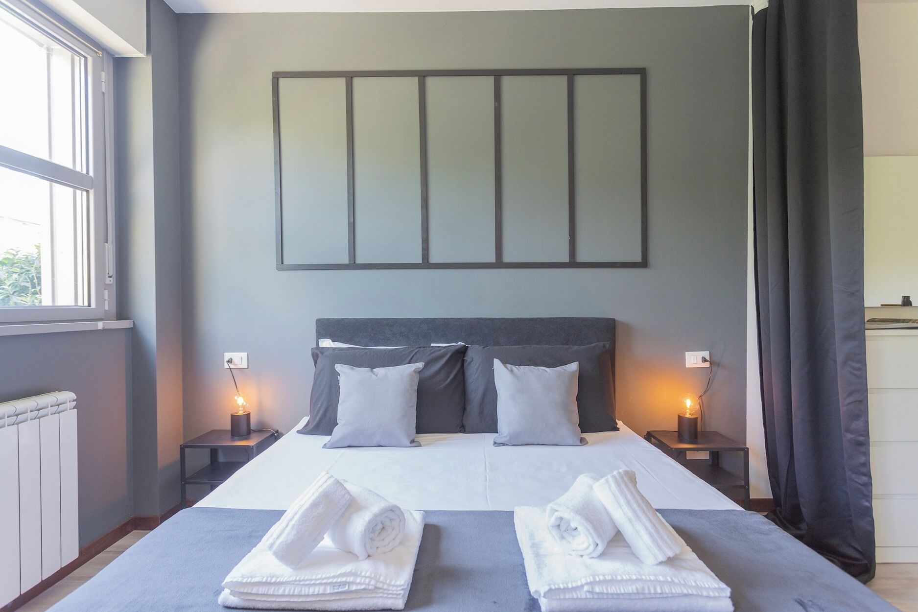 Ein modernes Hotelzimmer mit einem großen Bett, zwei Nachttischen, einem Wandbild und Blick ins Freie.