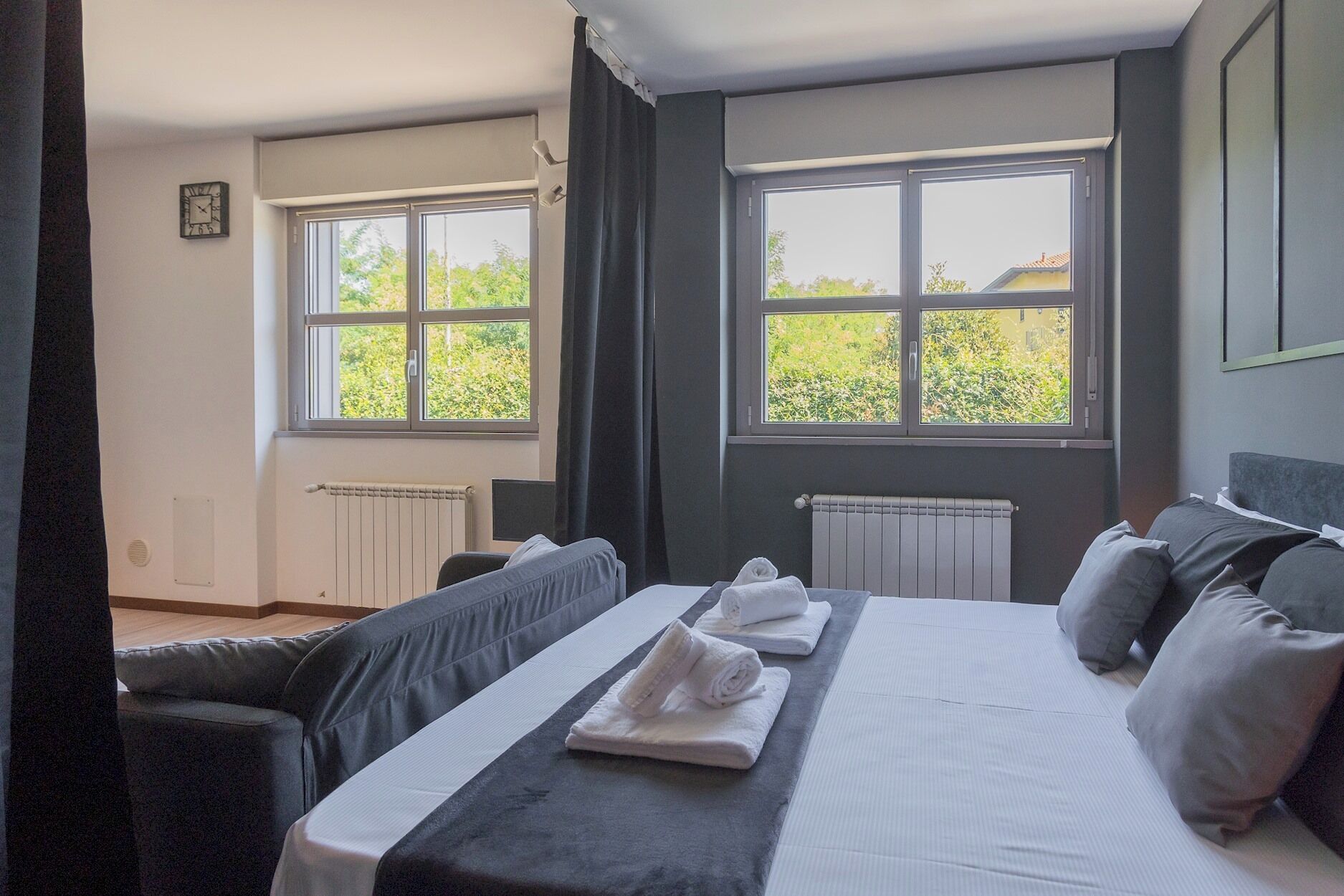 A hotel room with a bed, two windows, a radiator, and a view of greenery outside.