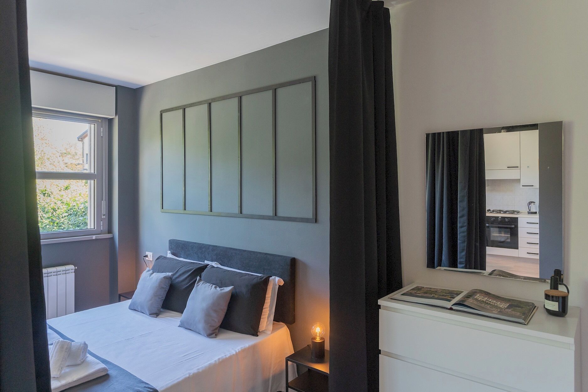Un dormitorio con cama, una mesita de noche con lámpara, un espejo grande y vistas a la cocina.