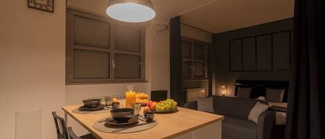 A modern kitchen with a dining table, chairs, and a pendant light. There is a fruit bowl on the table and a window with blinds.