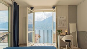 A bedroom with a bed, a desk, a chair, and a view of a lake and mountains.